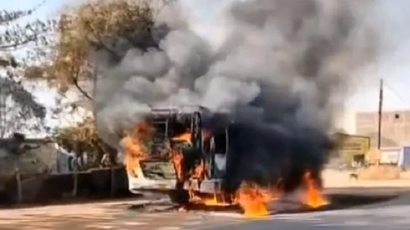 A passenger bus engulfed in massive orange flames and black smoke on a rural Chhattisgarh highway, police personnel in distance.