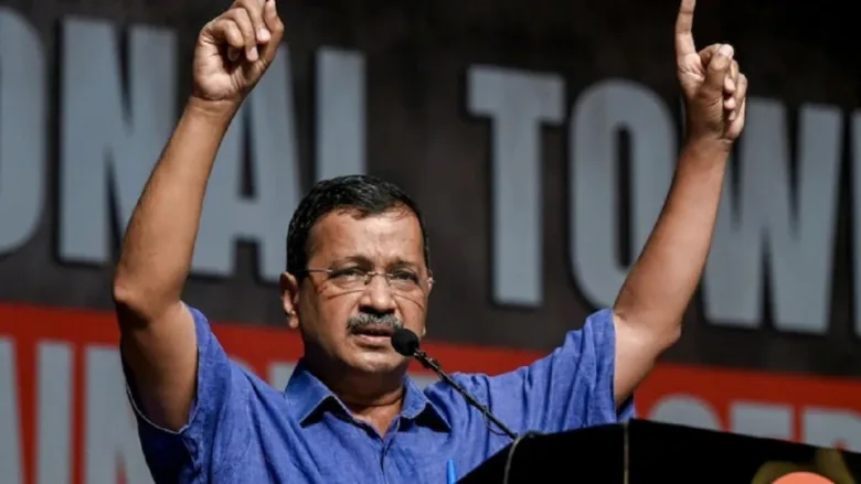 मंच से भाषण देते हुए अरविंद केजरीवाल, दोनों हाथ ऊपर उठाकर लोगों को संबोधित करते हुए।